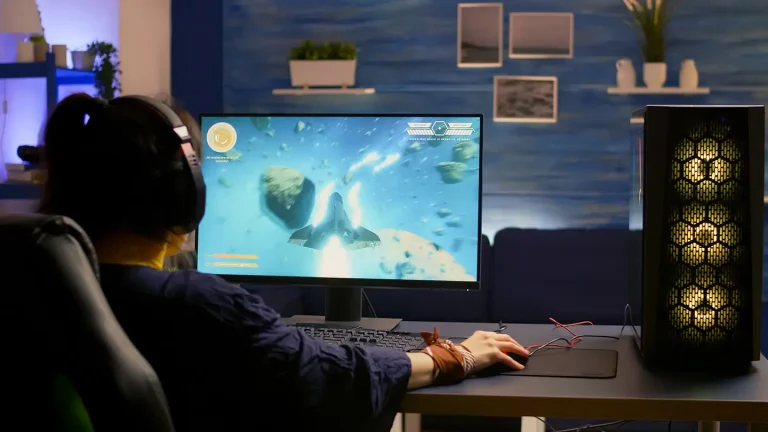 Gracz w słuchawkach siedzi przy biurku i gra na monitorze gamingowym z dużym ekranem; obok stoi komputer stacjonarny z podświetleniem RGB.