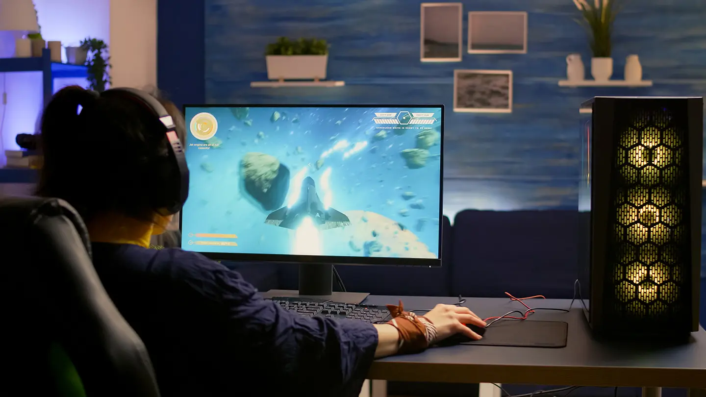 Gracz w słuchawkach siedzi przy biurku i gra na monitorze gamingowym z dużym ekranem; obok stoi komputer stacjonarny z podświetleniem RGB.